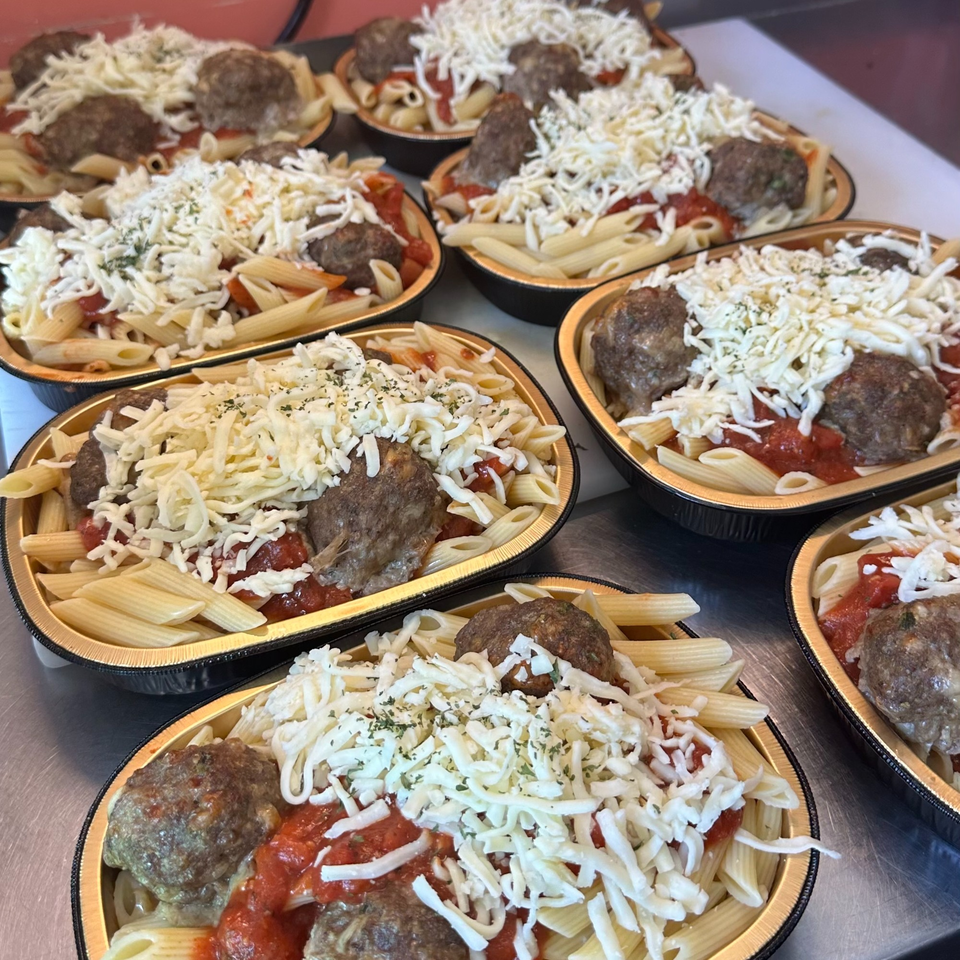 Multiple foil containers filled with penne pasta, meatballs, tomato sauce, and shredded cheese.