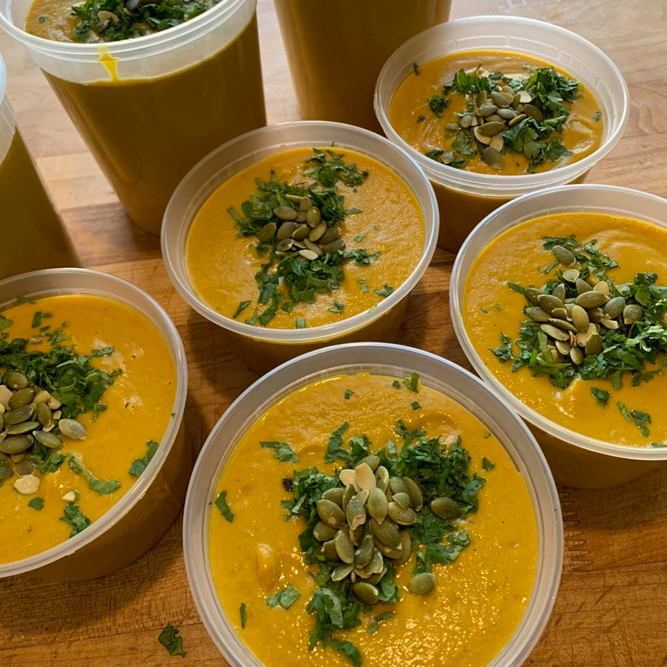 Several clear plastic containers filled with vibrant orange soup, garnished with green herbs and pumpkin seeds.