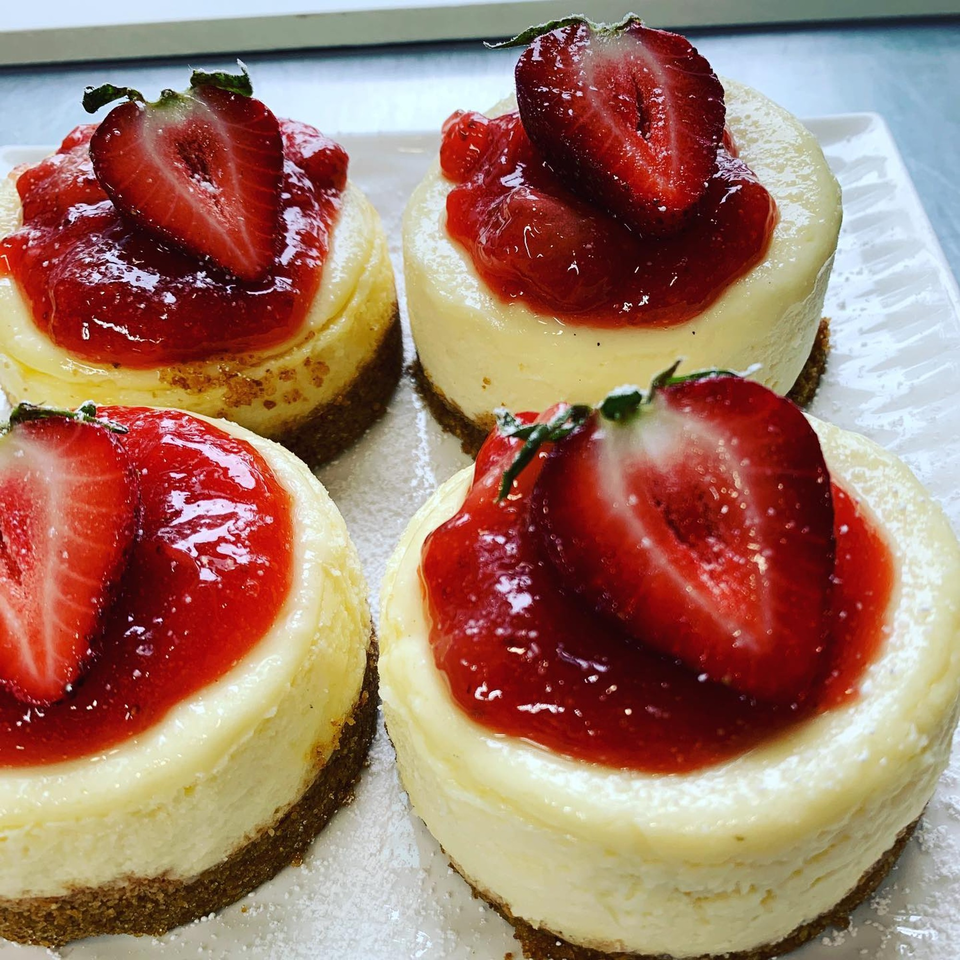 Four mini cheesecakes with graham cracker crusts, topped with strawberry compote and a fresh strawberry half.
