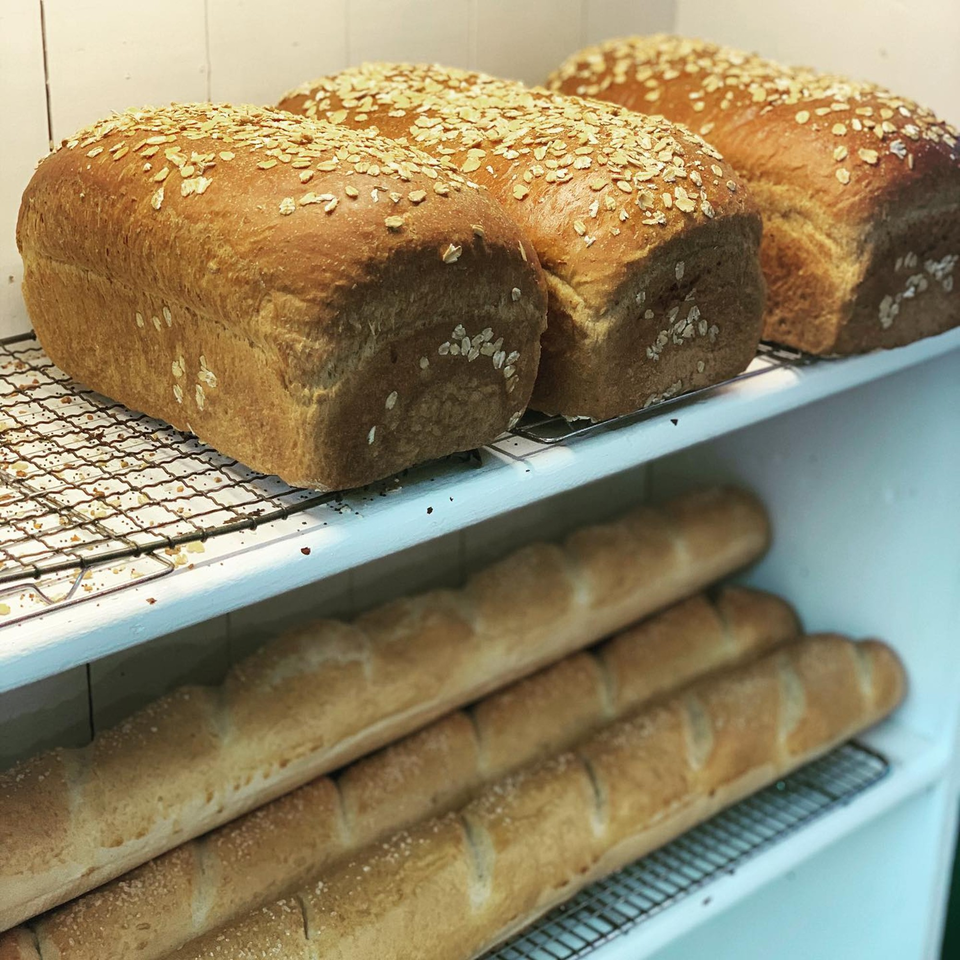 Freshly baked loaves of oat-topped bread and baguettes on cooling racks at Hello Good Pie bakery.